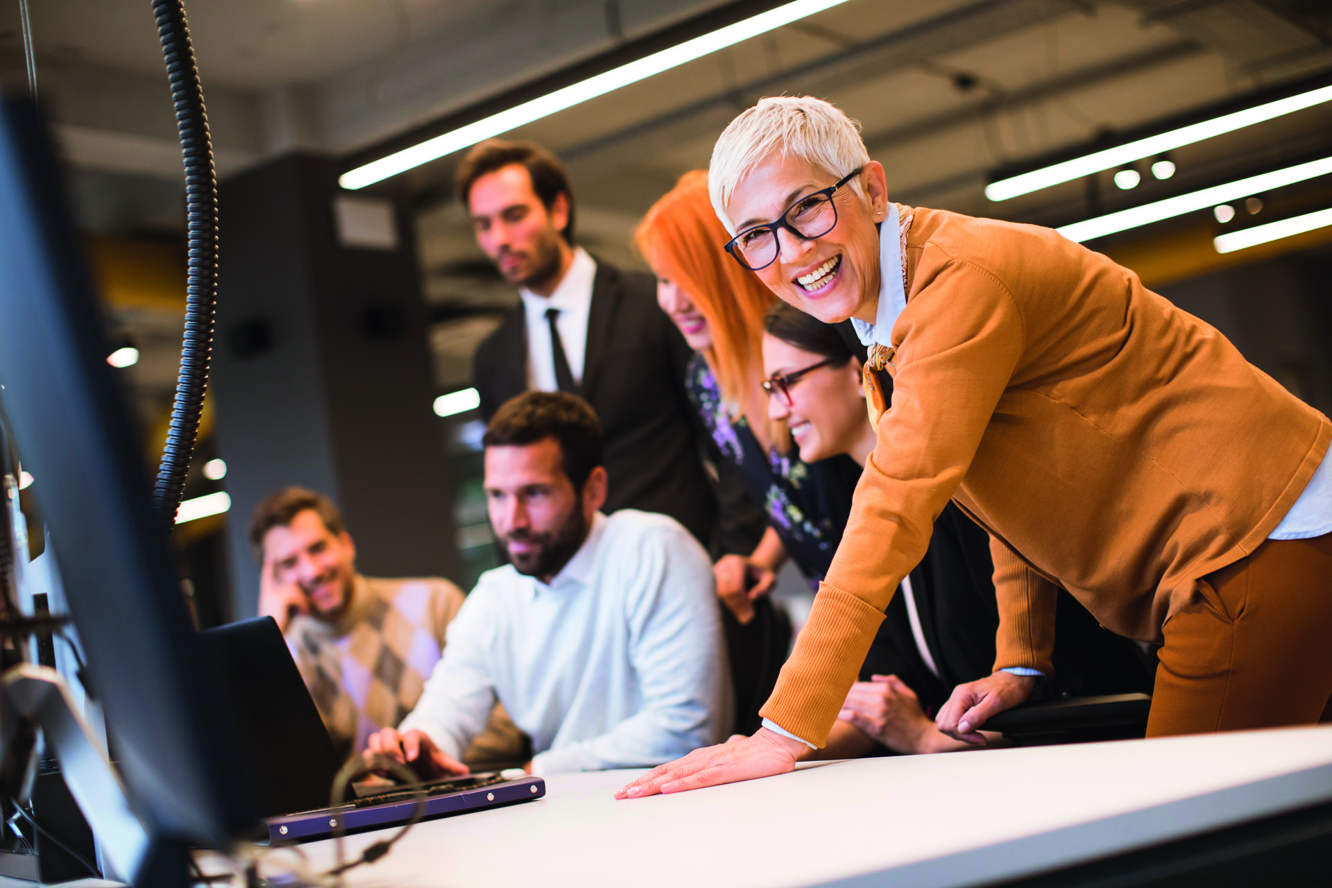 Dame, mit weißen Haaren und markanter schwarzer Brille schaut seitlich zum Betrachter und lehnt sich mit einer Hand am Schreibtisch.