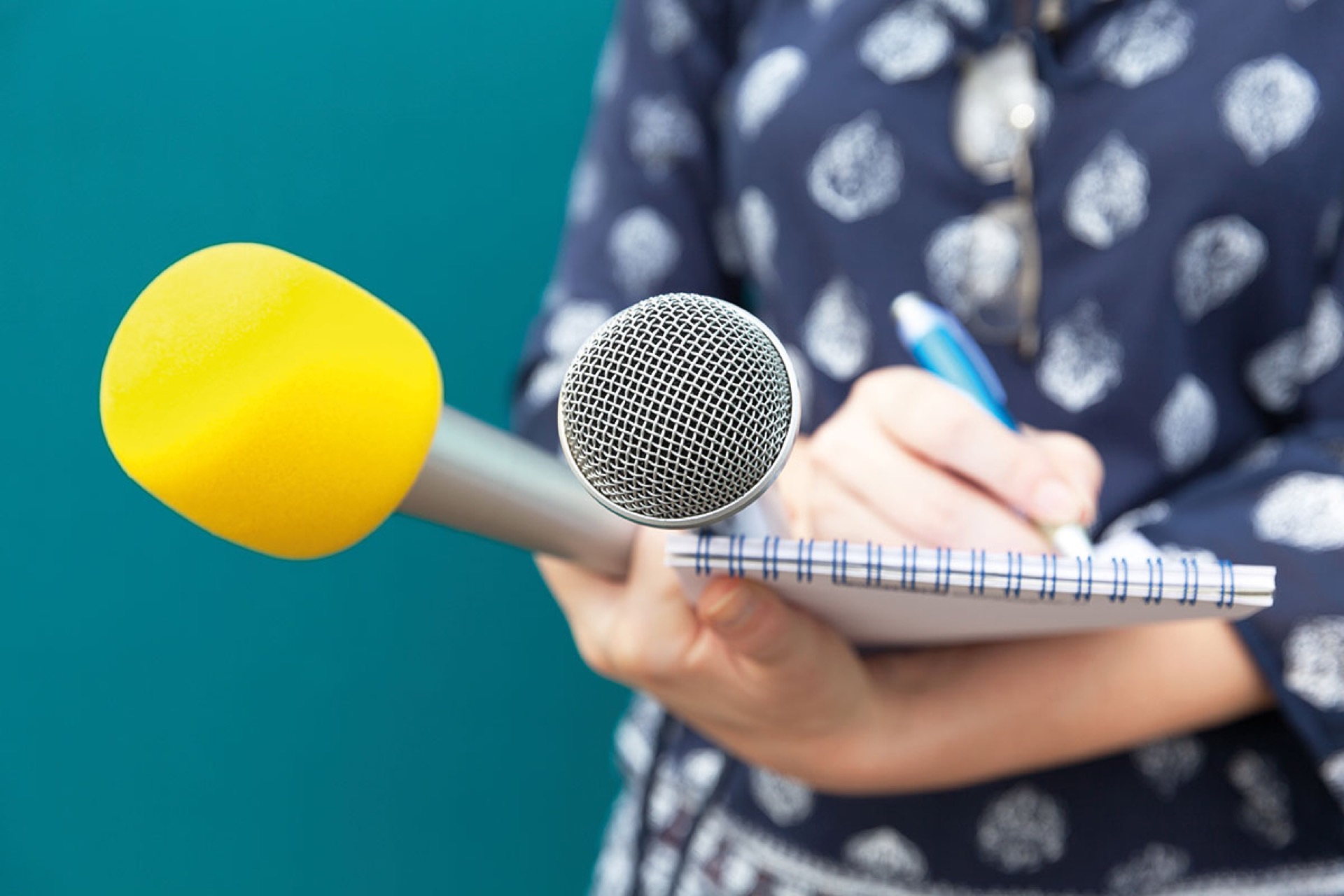 Frau in gepunktetem Kleid schreibt in einen Schreibblock, auf dem ein  gelbes und graues Mikrofon liegen.