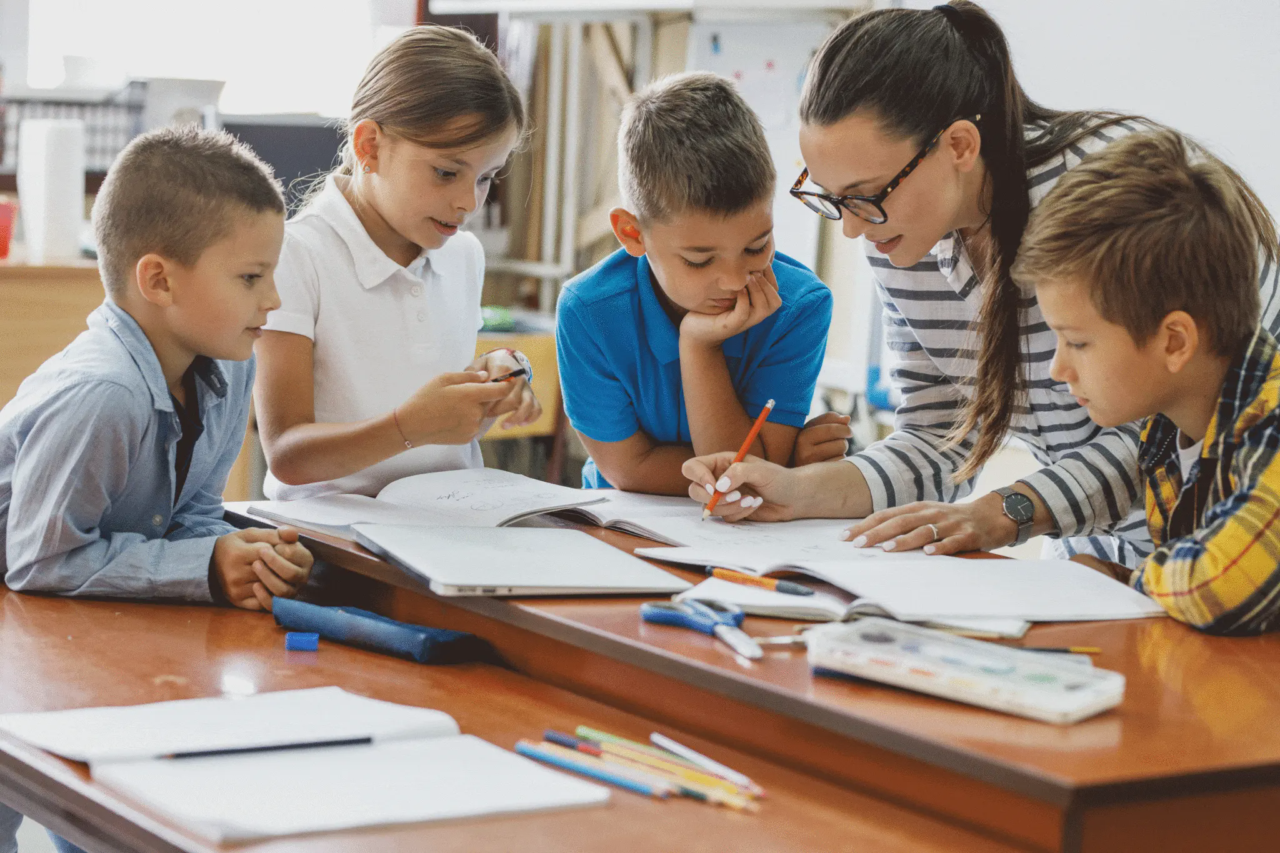 Lehrerin korrigiert vor den sie umzingelnden Schülern eine Aufgabe in den Heften der Kinder.
