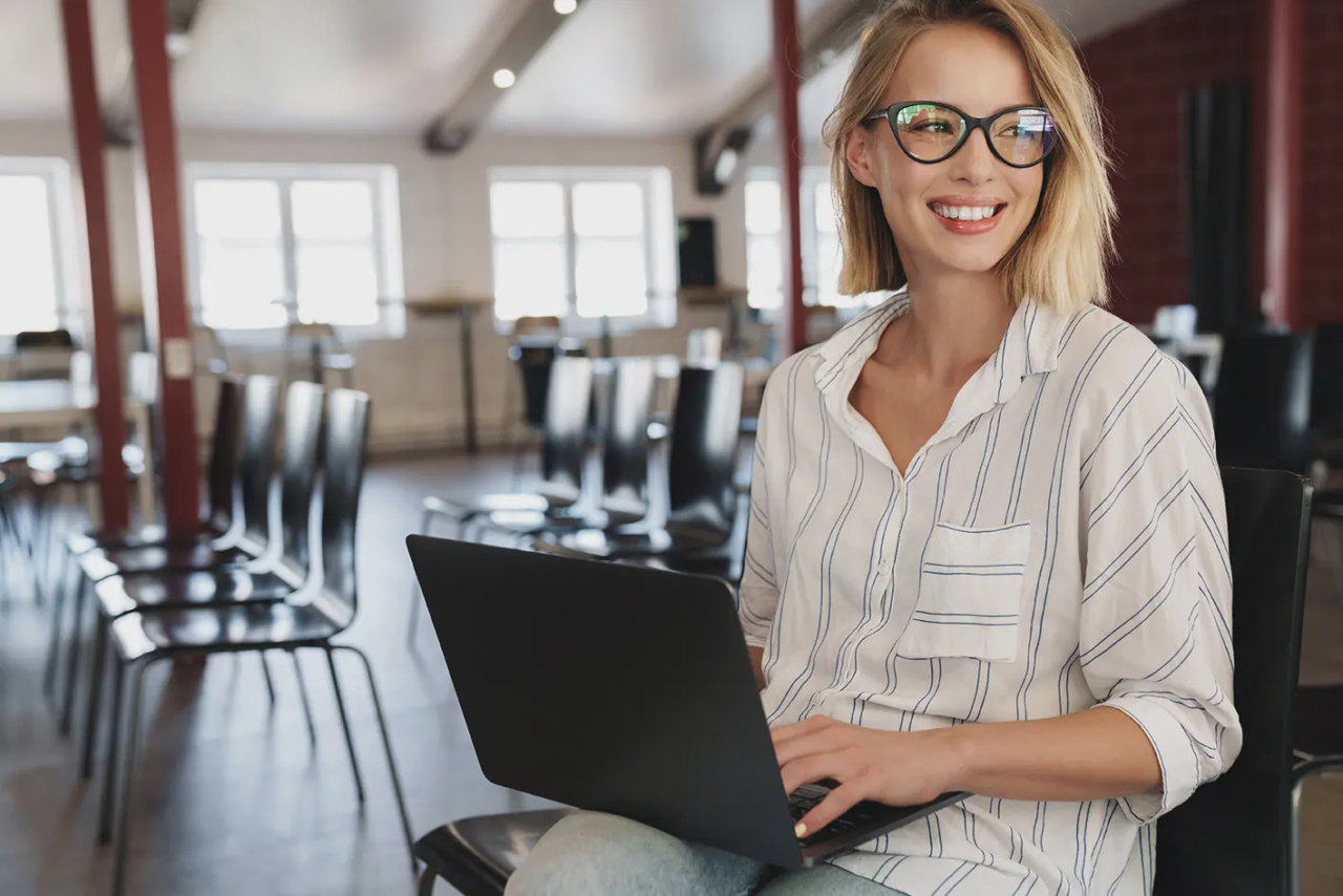 Blonde Frau mit schwarzer, markanter Brille sitzt auf einem Stuhl mit einem Laptop auf dem Schoß in einem großen Raum und schaut rechts schräg aus dem Bild raus.