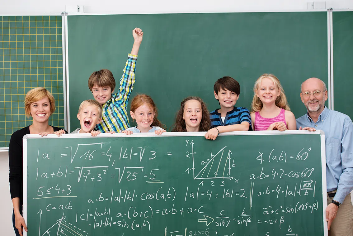 Eine Lehrerin und ein Lehrer stehen mit den Grundschülern vor der Schultafel und halten selbst auch eine große Tafel vor sich mit komplizierten mathematischen Ausdrücken.